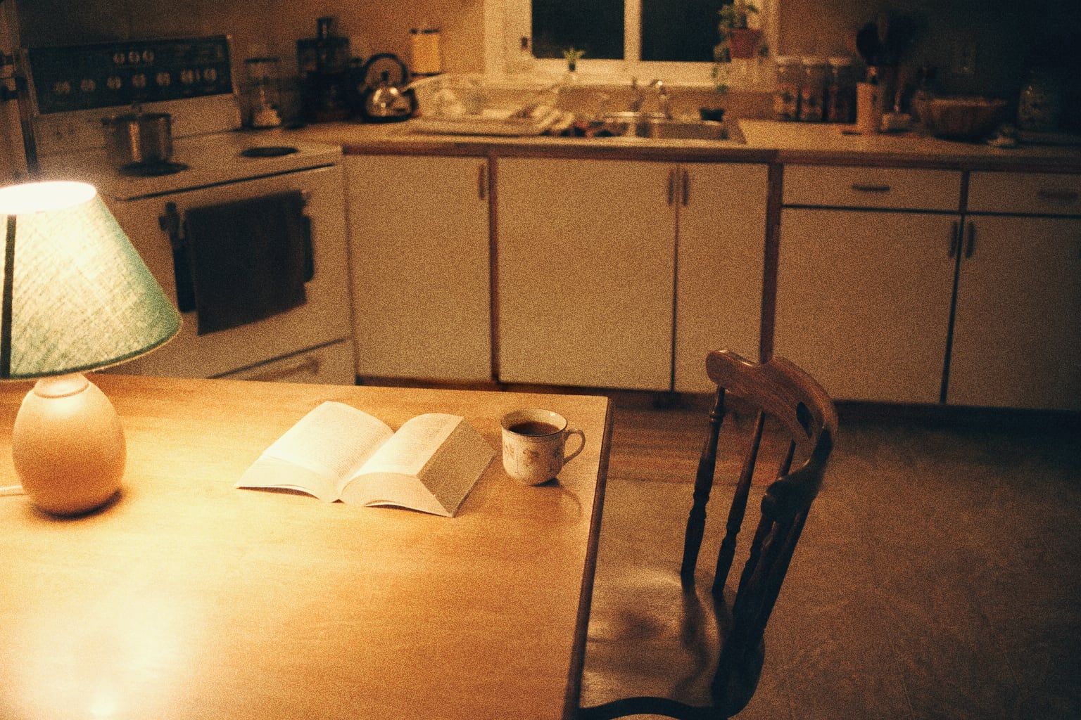 A 1990s kitchen at night in warm lamplight, an open paperback and a cup of tea on the table, one wooden chair pulled out, shot on grainy film.